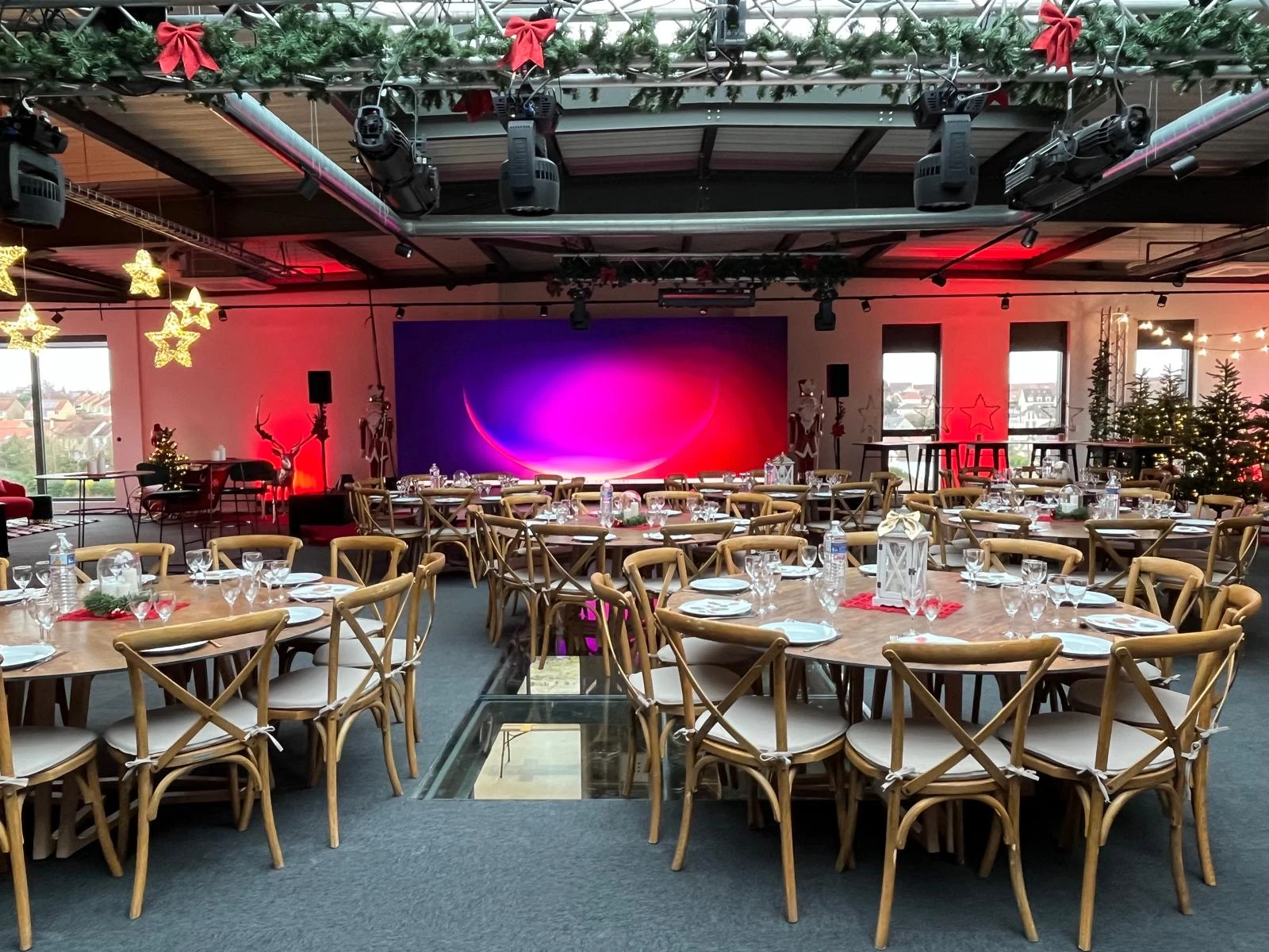 Salle de réception décorée pour Noël avec tables rondes, chaises en bois, étoiles lumineuses et sapins illuminés